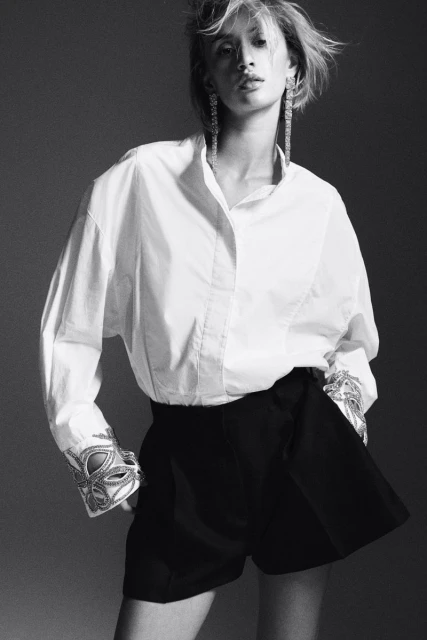Fashion shot of a woman wearing a white shirt and black shorts, accessorized with long earrings and decorative cuffs in a grey studio.