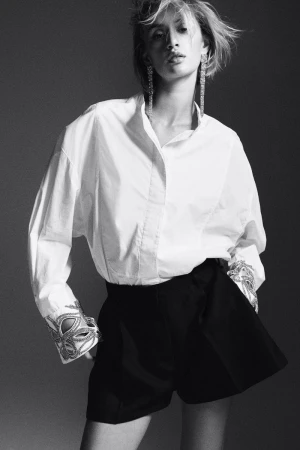 Fashion shot of a woman wearing a white shirt and black shorts, accessorized with long earrings and decorative cuffs in a grey studio.