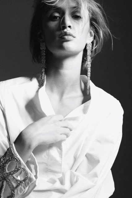 Black-and-white portrait of a fair-skinned adult female wearing long dangling earrings and an unbuttoned white shirt in a studio.