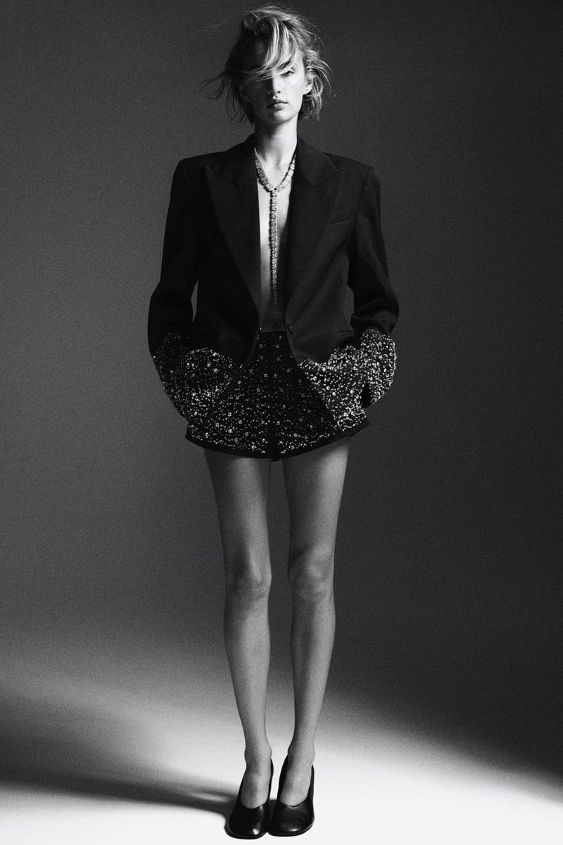Full-length studio shot of a blonde female model in a black blazer, embellished shorts, and heels, striking a pose against a grey background.