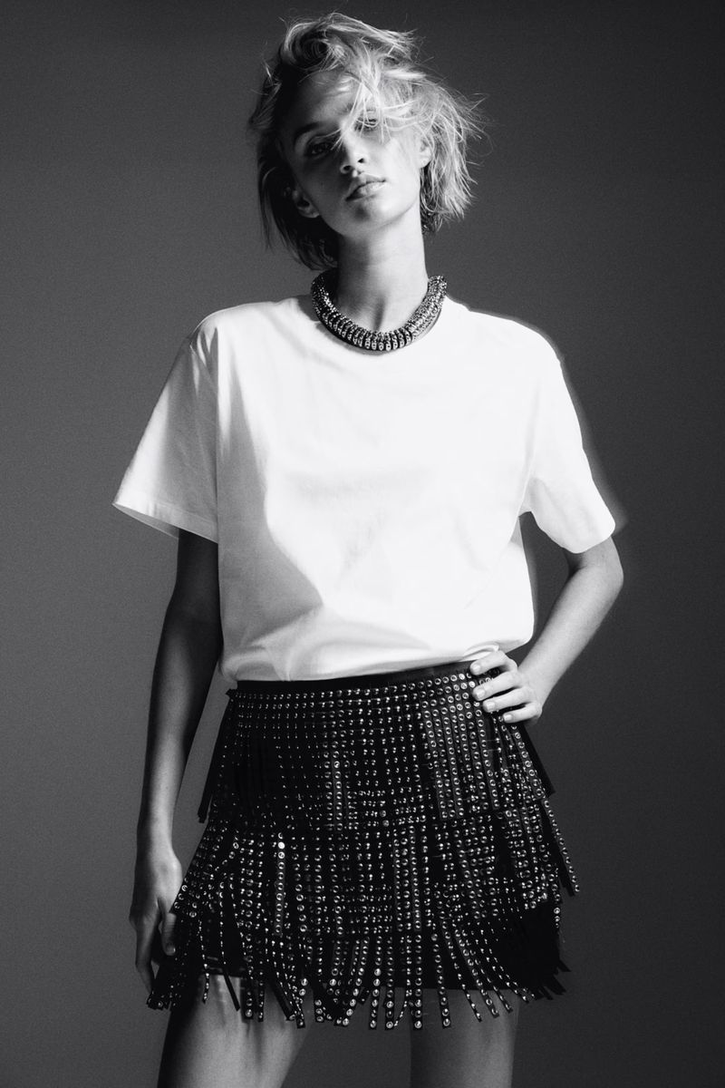 Black and white portrait of a woman in a white T-shirt and beaded skirt with hand on hip in a studio setting with a necklace.
