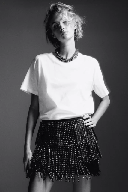 Black and white portrait of a woman in a white T-shirt and beaded skirt with hand on hip in a studio setting with a necklace.