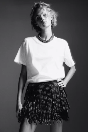 Black and white portrait of a woman in a white T-shirt and beaded skirt with hand on hip in a studio setting with a necklace.
