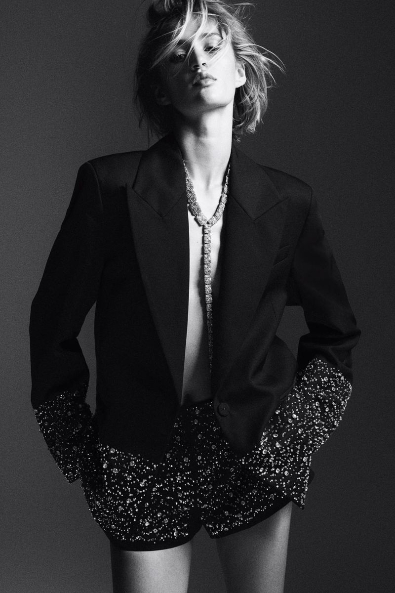 Monochrome fashion shot: woman poses in blazer, shorts, and necklace against a grey backdrop, hands in pockets.