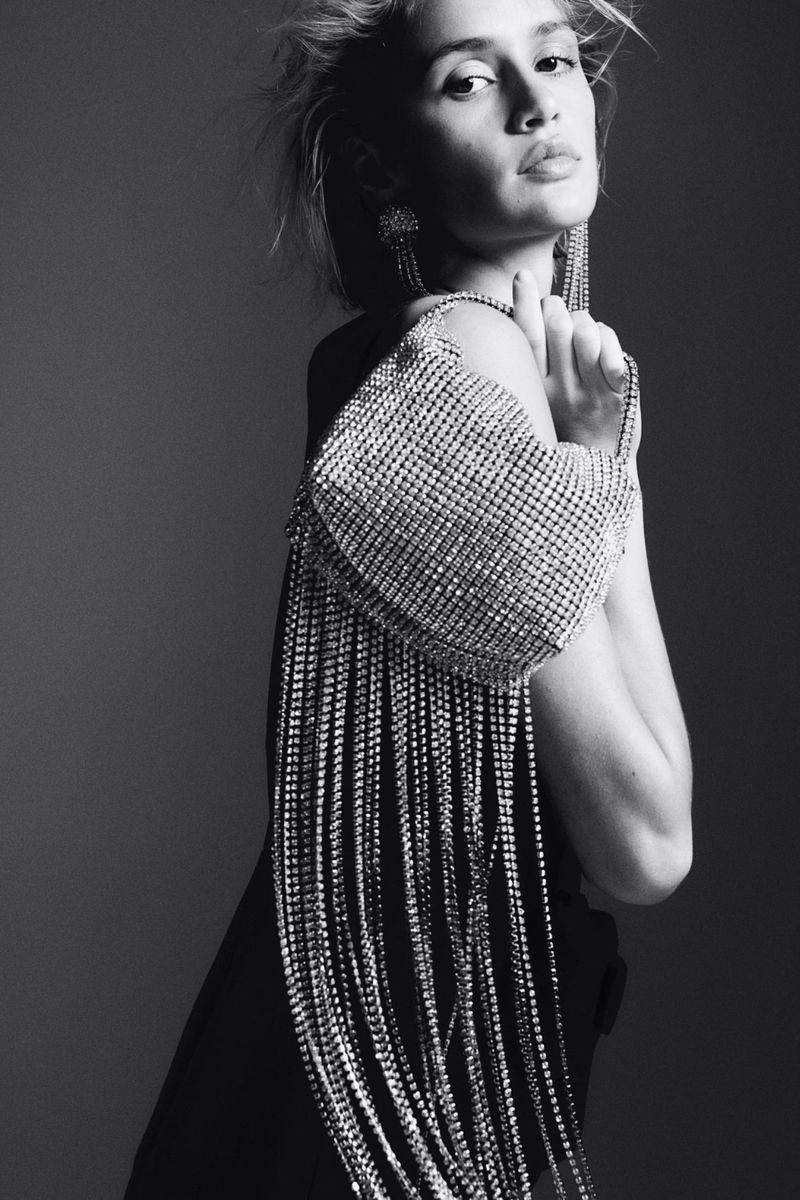 Monochrome portrait of a woman with a rhinestone fringe purse, statement earrings, and hand near her face in a studio setting.