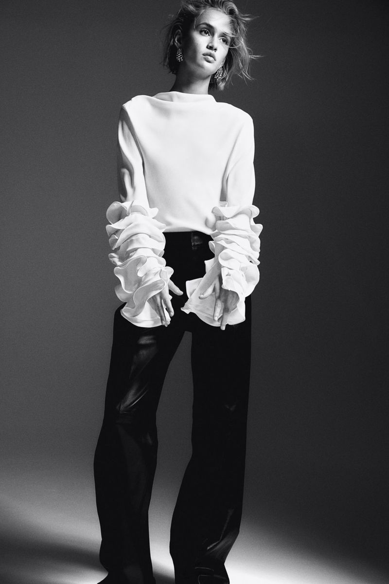 Model in white ruffled blouse and dark trousers against a neutral studio background for H&M Holiday 2024 lookbook