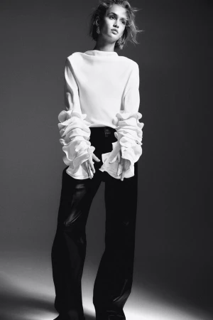 Model in white ruffled blouse and dark trousers against a neutral studio background for H&M Holiday 2024 lookbook