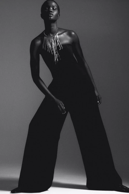 Black and white studio portrait of a Black female model in wide-leg jumpsuit with statement necklace posing against a neutral backdrop.