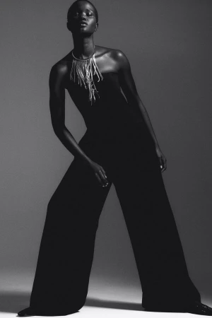 Black and white studio portrait of a Black female model in wide-leg jumpsuit with statement necklace posing against a neutral backdrop.