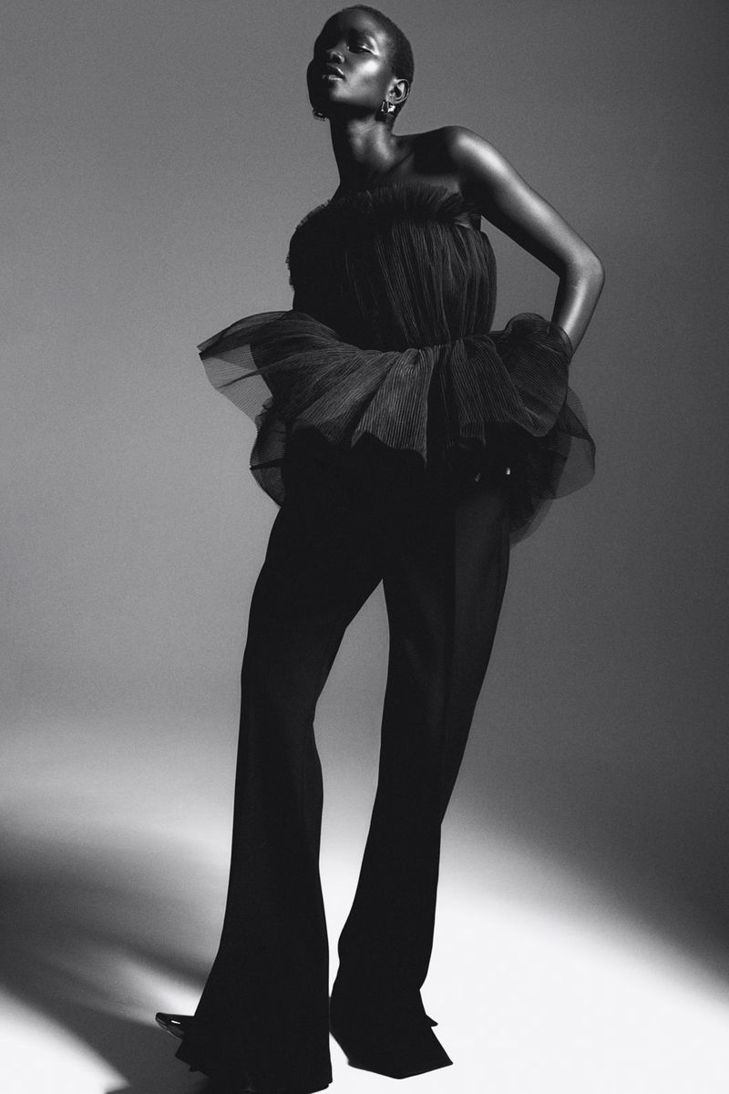 Black and white studio shot of an African adult female model in a black tulle top and wide-leg pants, posing with a serious expression.