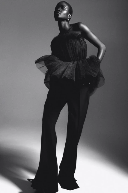 Black and white studio shot of an African adult female model in a black tulle top and wide-leg pants, posing with a serious expression.