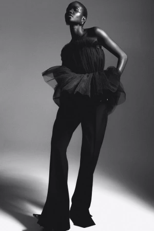 Black and white studio shot of an African adult female model in a black tulle top and wide-leg pants, posing with a serious expression.