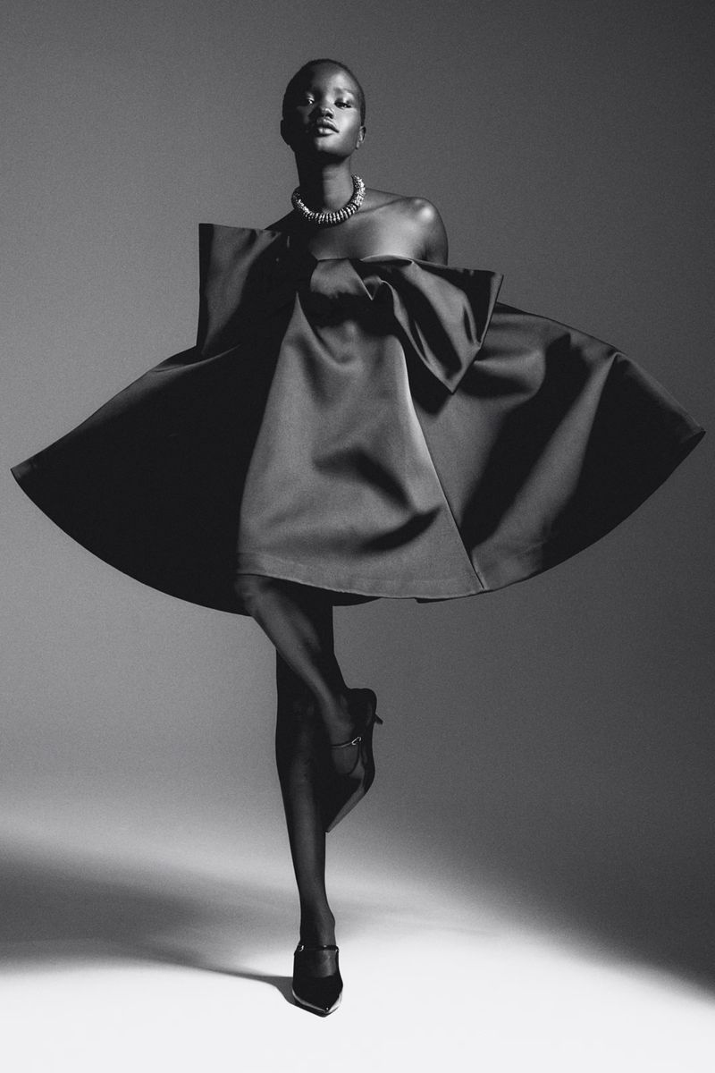 Elegant black and white fashion portrait of a woman in a satin dress, accessorized with a necklace and heels, against a neutral studio backdrop.