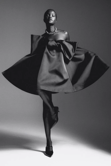 Elegant black and white fashion portrait of a woman in a satin dress, accessorized with a necklace and heels, against a neutral studio backdrop.