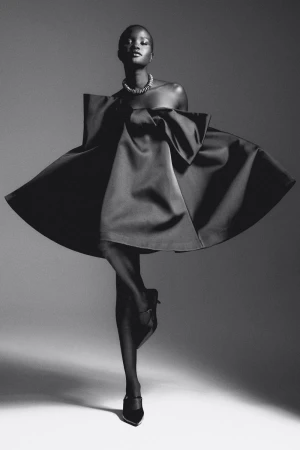 Elegant black and white fashion portrait of a woman in a satin dress, accessorized with a necklace and heels, against a neutral studio backdrop.