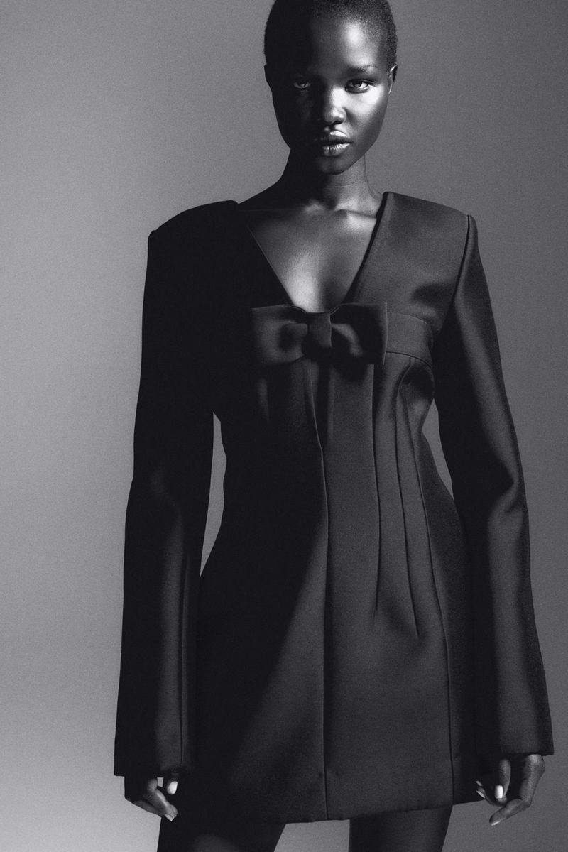 Glamorous Black and white studio portrait of a Black woman in a dark dress with a bow, posing against a gradient gray backdrop.