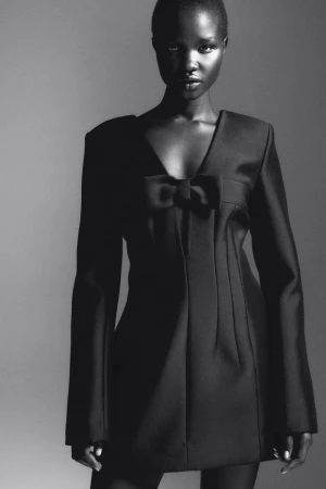 Glamorous Black and white studio portrait of a Black woman in a dark dress with a bow, posing against a gradient gray backdrop.
