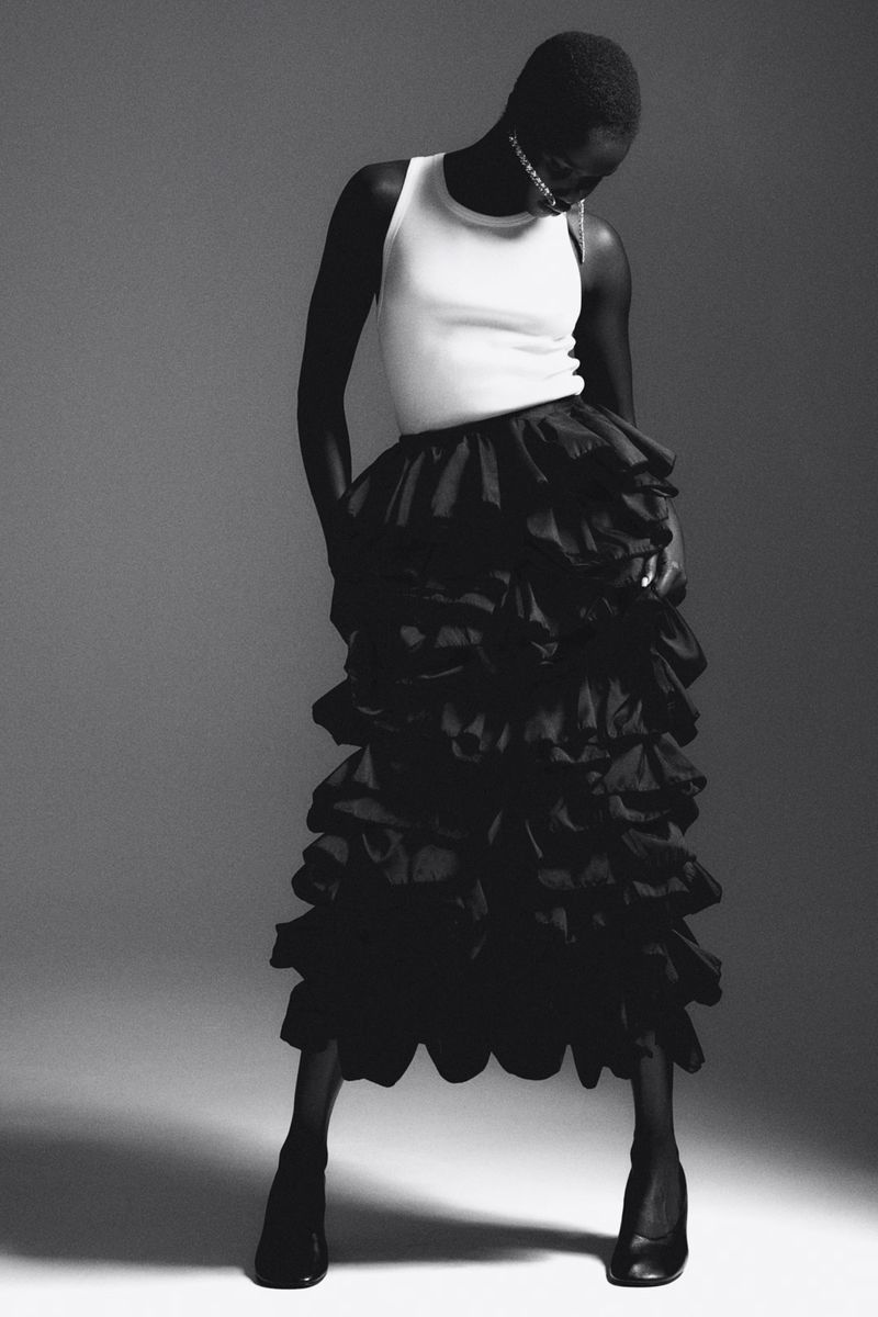 Editorial fashion shot of a Black female model wearing a white tank top and black ruffled skirt in a studio with grey backdrop.