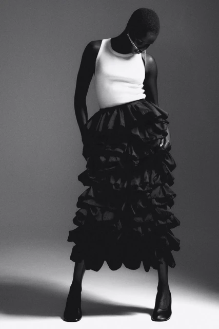 Editorial fashion shot of a Black female model wearing a white tank top and black ruffled skirt in a studio with grey backdrop.