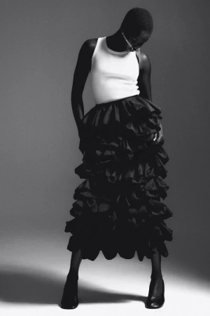 Editorial fashion shot of a Black female model wearing a white tank top and black ruffled skirt in a studio with grey backdrop.