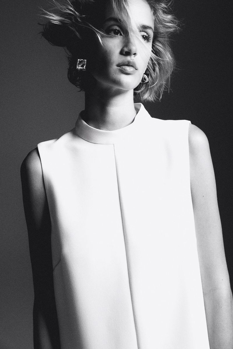 Black and white portrait of a fair-skinned woman with short hair, wearing a white sleeveless dress and statement earrings, in studio lighting.