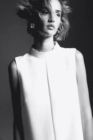 Black and white portrait of a fair-skinned woman with short hair, wearing a white sleeveless dress and statement earrings, in studio lighting.