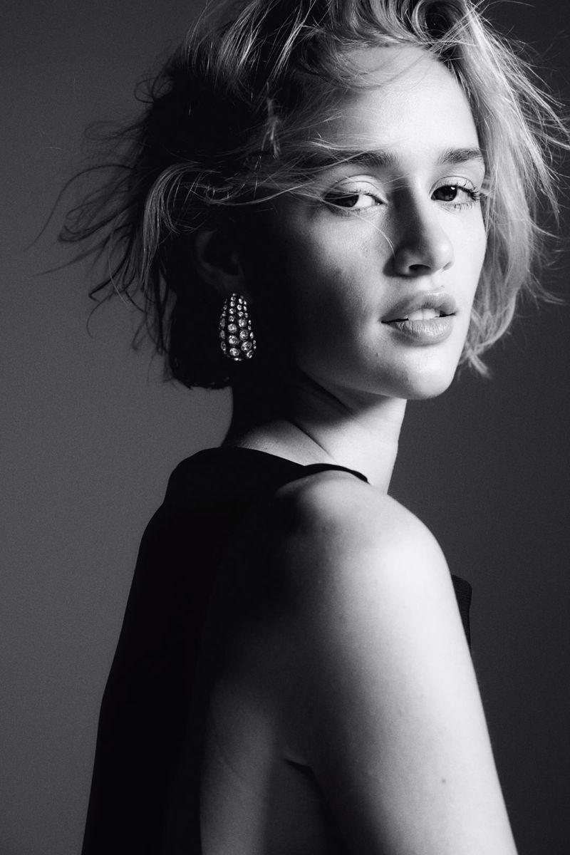 Black and white portrait of a woman with short tousled hair wearing statement earrings and a dark top against a grey studio backdrop.