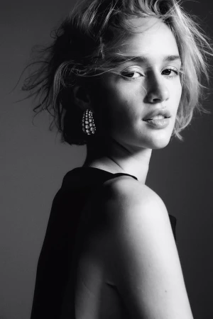 Black and white portrait of a woman with short tousled hair wearing statement earrings and a dark top against a grey studio backdrop.