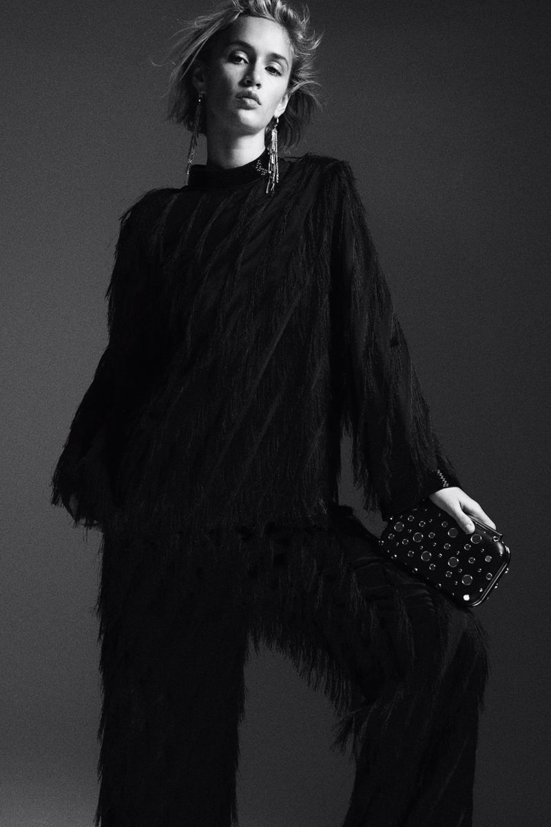 Elegant woman in black fringe top and pants holds studded clutch against a muted grey backdrop in a studio fashion shot.