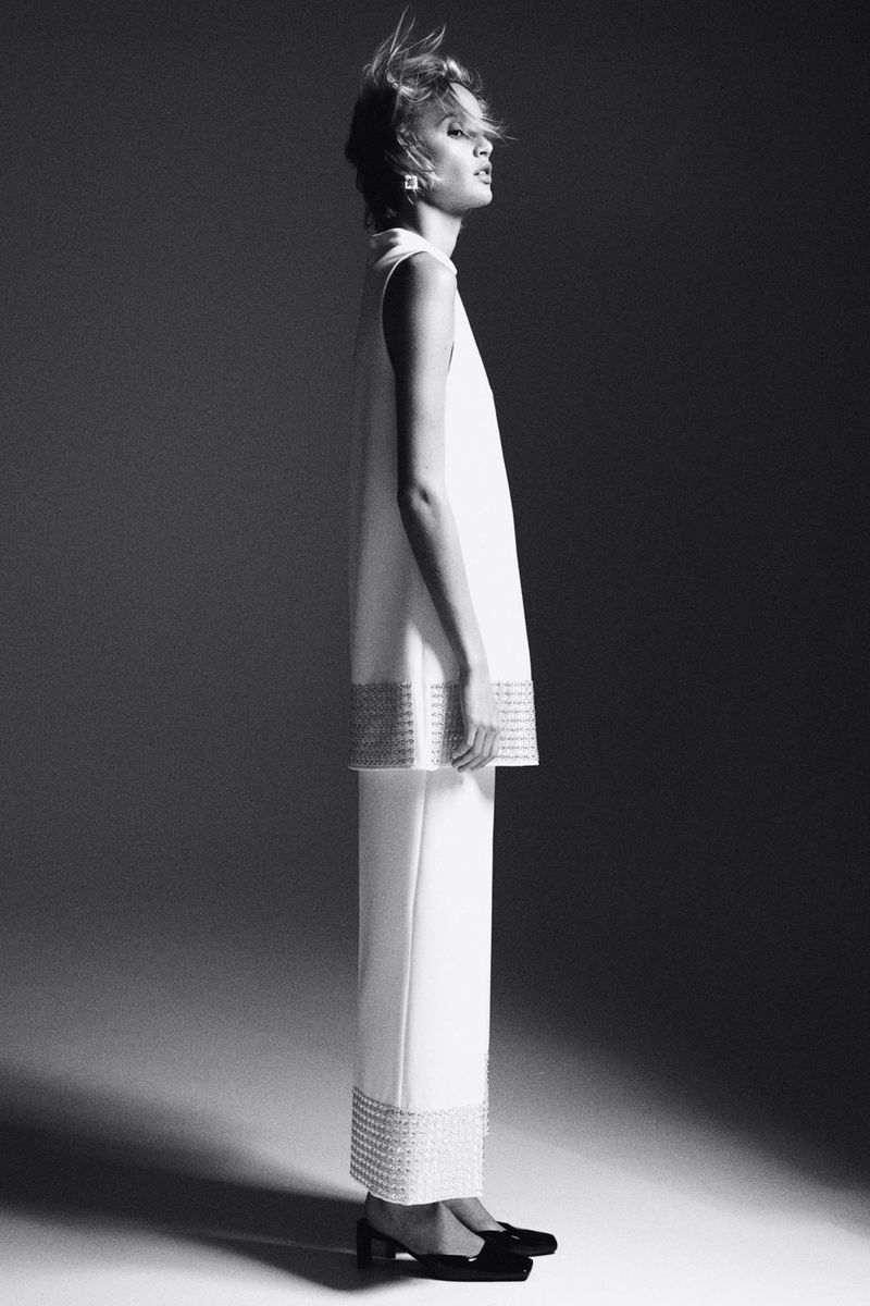 Monochrome studio shot of a fair-skinned adult female in a white, sleeveless dress and pants, standing in profile with a neutral expression.