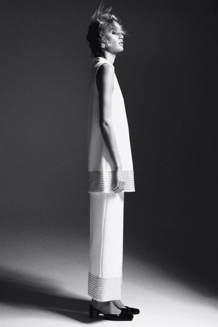 Monochrome studio shot of a fair-skinned adult female in a white, sleeveless dress and pants, standing in profile with a neutral expression.