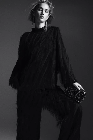Model in black fringe top and trousers holding a studded clutch against a grey studio backdrop