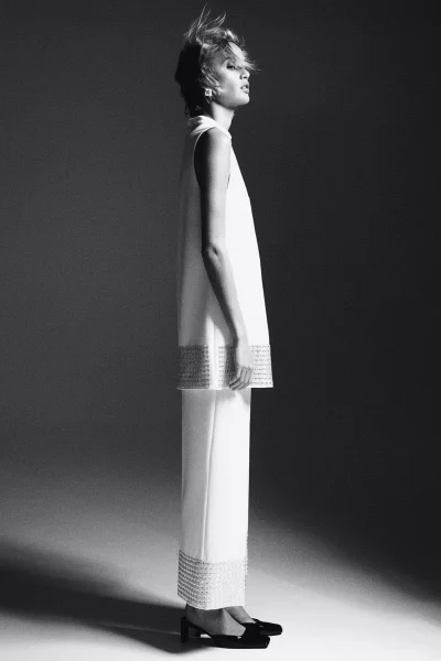 Model in a white sleeveless top and trousers standing in profile against a dark gradient studio background