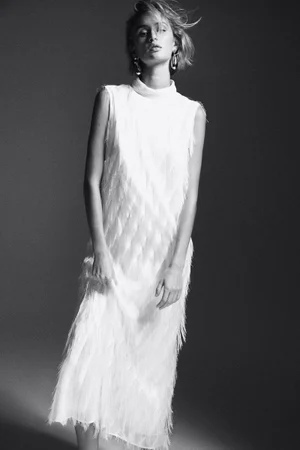 Model wearing a white fringed sleeveless dress with statement earrings in a monochrome studio lookbook image