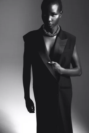 Black and white studio portrait of a model wearing a tailored sleeveless blazer and a thick textured statement necklace.