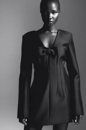 Model in a black mini dress featuring structured shoulders and a central bow accent against a seamless gradient background.