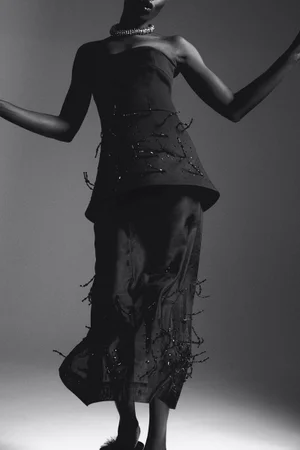Model in a black strapless dress adorned with beaded fringe, wearing a statement necklace against a grey seamless background