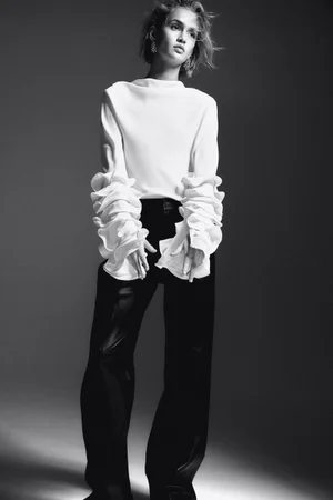 Model in white ruffled blouse and dark trousers against a neutral studio background for H&M Holiday 2024 lookbook