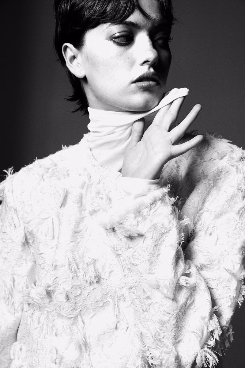Monochrome portrait of a woman with short dark hair, wearing a white, textured top and a white scarf, against a grey background.