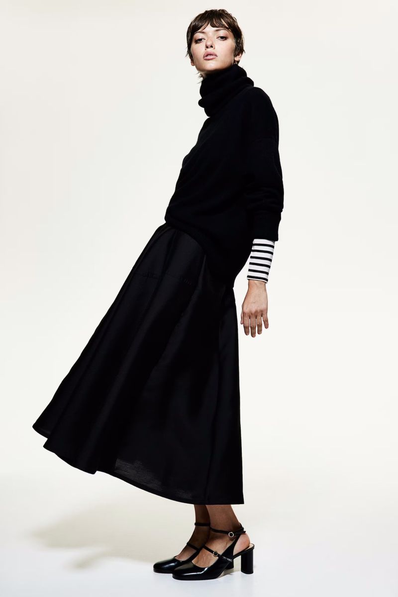 Fashion model in black turtleneck sweater and skirt stands in a studio setting, showcasing a stylish, elegant, and monochromatic look.