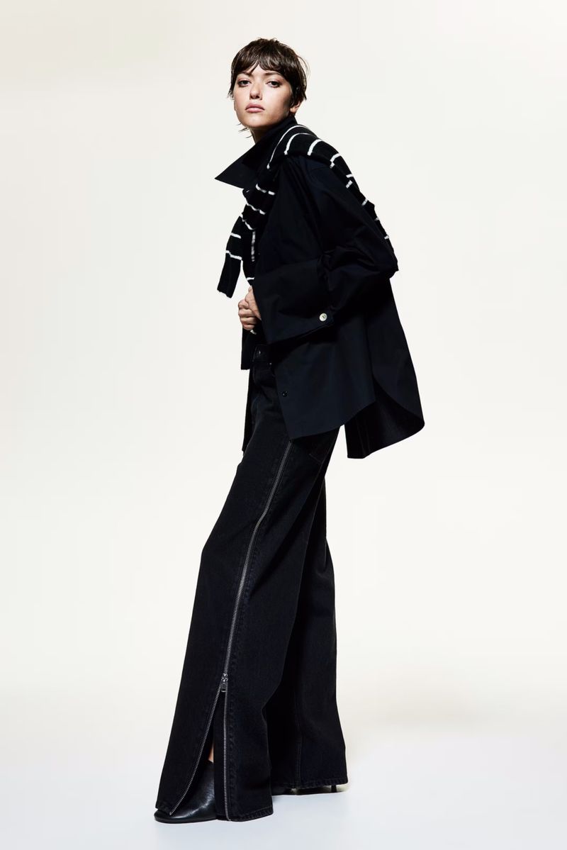 Woman in black coat and flared pants strikes a confident pose against a bright white backdrop, showcasing fashion and style.
