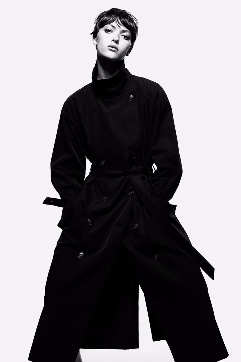 A slender adult female model with short brown hair poses indoors in a black, belted trench coat with a white background in a fashion shot.