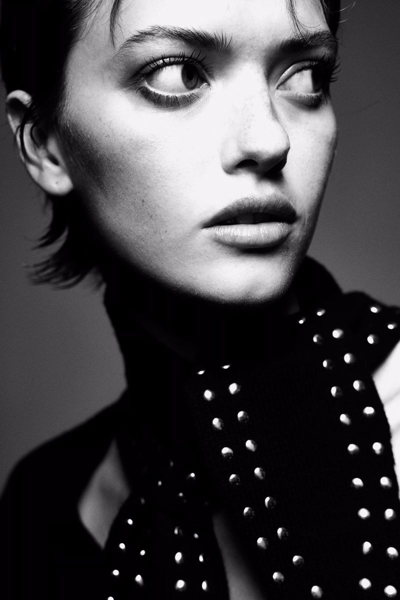 Black and white portrait of a young adult woman with short hair looking to the side, wearing a studded shirt and a vest.