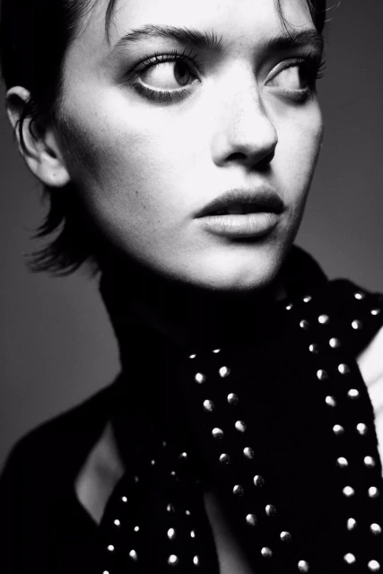 Black and white portrait of a young adult woman with short hair looking to the side, wearing a studded shirt and a vest.