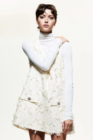 Model with short dark hair wearing a white turtleneck and textured cream dress against a bright white background