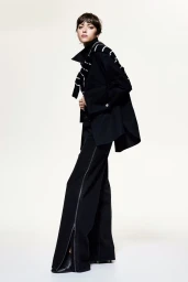 Model in black jacket and trousers with a striped sweater draped over shoulders against a plain white background