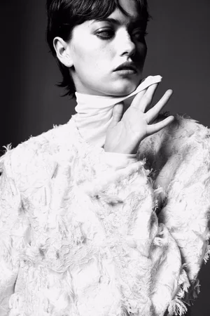 Model with dark short hair wearing a white textured fringed top and high neck collar against a dark studio background