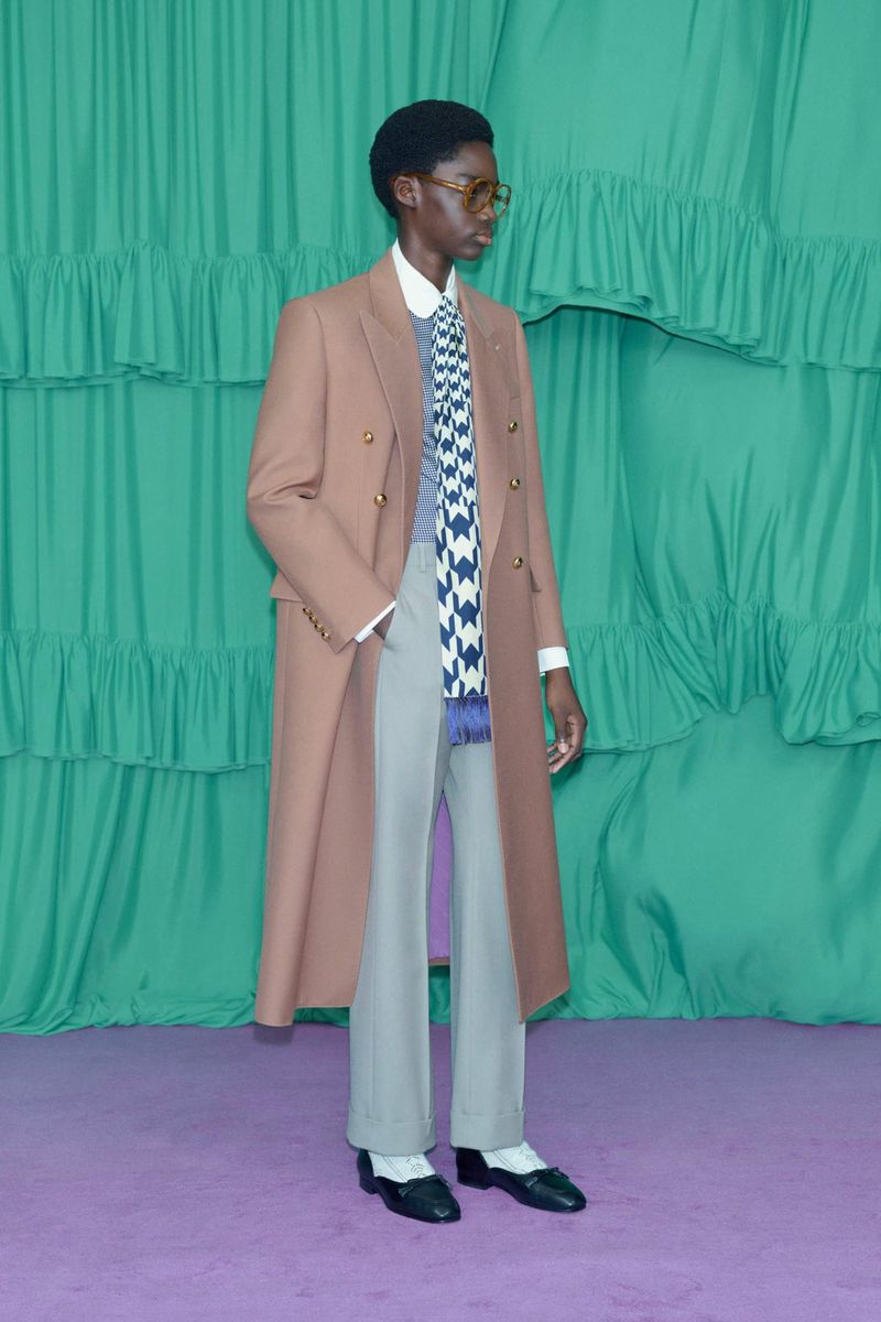 Full-length shot of an African-American male model wearing a tan coat, grey pants, scarf, and black loafers, against a green backdrop.