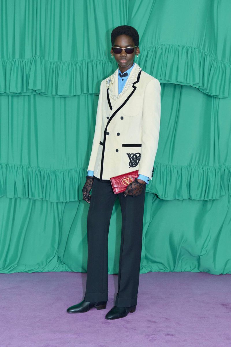 Fashionable Black man in double-breasted blazer with accessories stands on purple floor with green ruffled fabric backdrop.