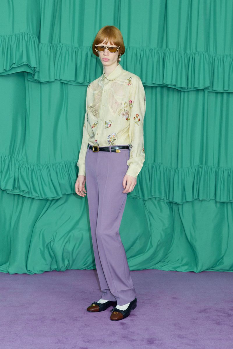 Adult fashion model poses in a semi-sheer button-down shirt, belted slacks, and loafers against a turquoise draped fabric background.